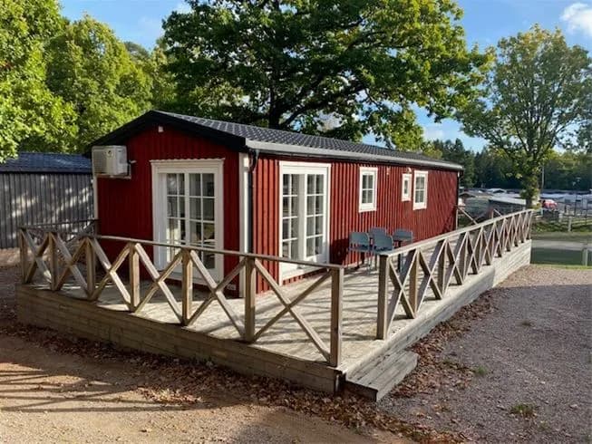 En pittoresk stuga vid Ursand Resort & Camping, komplett med en träveranda och omgivande grönska, erbjuder en inbjudande utomhusmiljö för besökare.