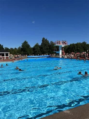 En bild av Skantzö Bad & Camping som visar glada besökare i och runt en utomhuspool, omgivna av gröna områden och campingfaciliteter. Besökare syns njuta av fritid och semesteraktiviteter i en bad- och campingmiljö.