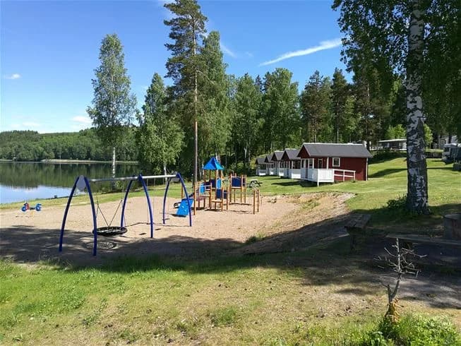 Bild av Degernäs camping som visar en grönskande parkmiljö med picknickbord och bänkar belägna i skuggan. En gräsmatta sträcker sig ner mot en sjö som utgör en del av landskapet, vilket ger en inbjudande plats för avkoppling och njutning av naturen.