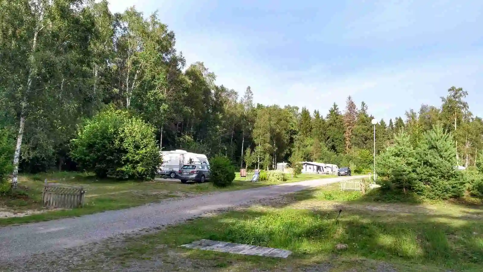 En campingplats omgiven av träd och växtlighet med flera parkerade husvagnar och bilar. I förgrunden syns en SUV och en mindre bil med registreringsskyltar. Området är inramat av barrträd.