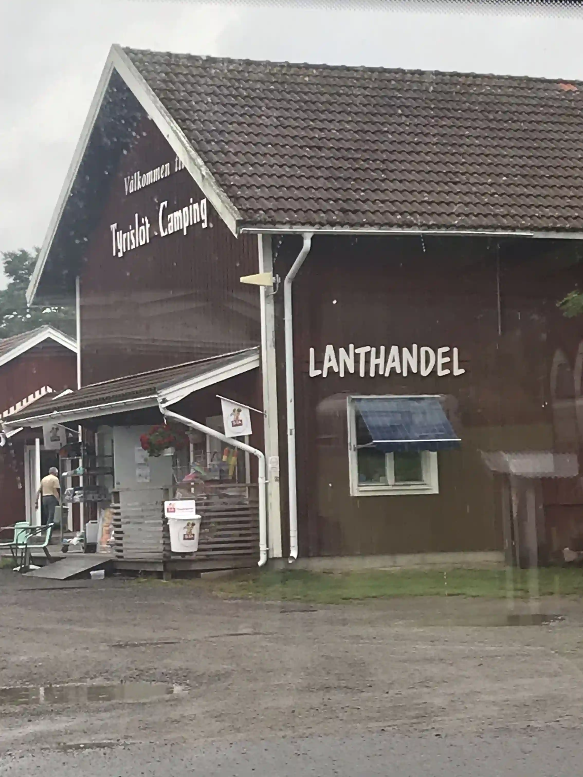 Bild av Tyrislöt Camping med ett färgglatt campingområde, där besökare kan se flera husvagnar och tält belägna nära vattnet. En solig dag med blå himmel, omgiven av frodig grönska. Perfekt för familjer och naturälskare.