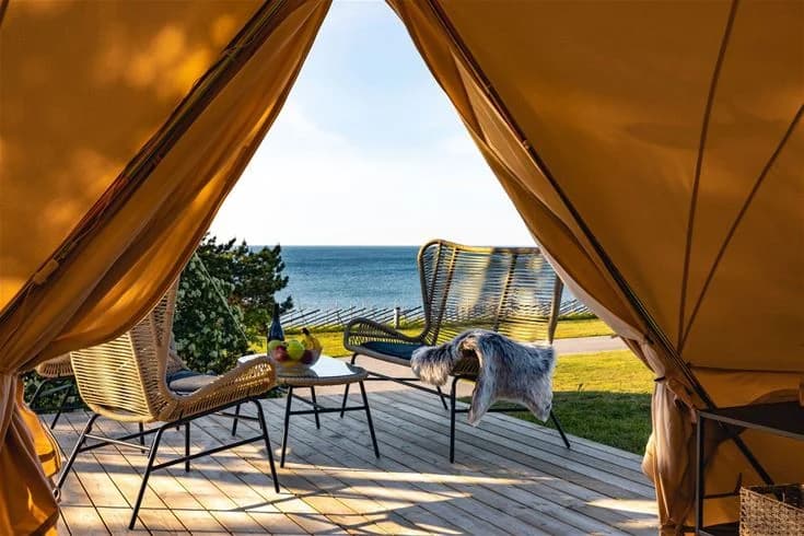 En strandnära vy vid Kneippbyn Resort Visby med havet och kustlinjen i bakgrunden. Soligt väder med skuggor från solens strålar. Utomhusmöbler och en campingplats med tält syns längs stranden.