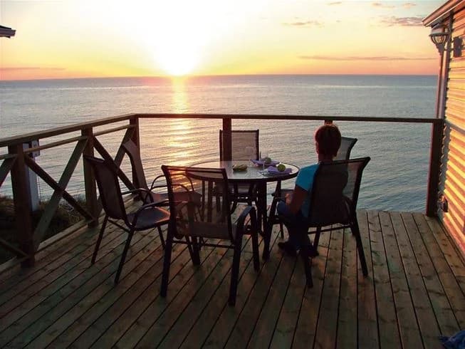 En solig sommardag vid Kneippbyn Resort Visby med utsikt över havet. I förgrunden syns en träbrygga och en balkong med utemöbler, medan horisonten sträcker sig ut över det glittrande vattnet.