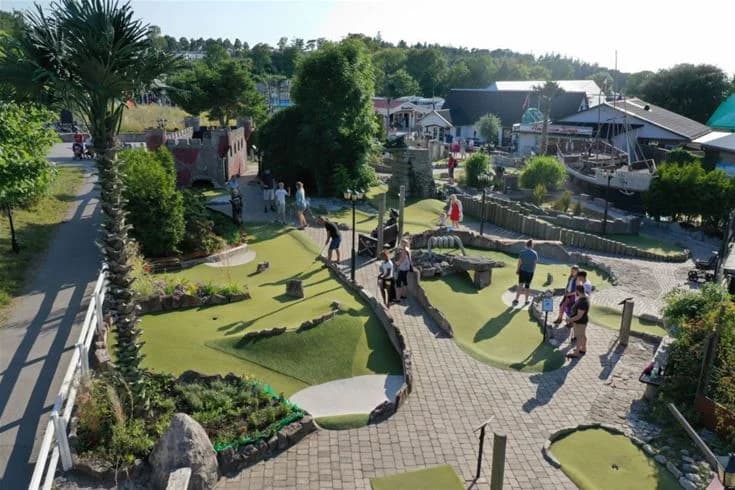 En översikt över Kneippbyn Resort Visby, som visar en vacker offentlig park med grönskande trädgårdar och anlagda områden. I förgrunden syns en minigolfbana där människor njuter av rekreation och avkoppling.