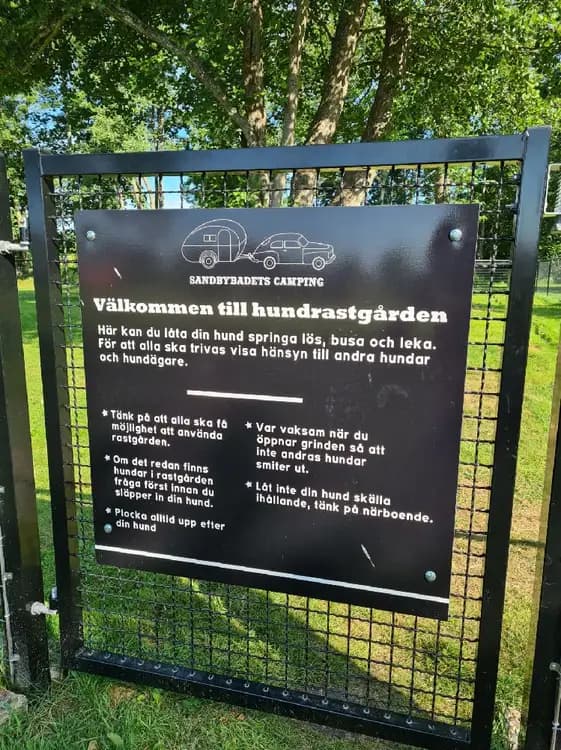 Alt-text: En grön campingplats vid Sandbybadet med en skylt och en minnesplakett omringad av gräs och markväxter, med en stig som leder genom ett naturskyddsområde.