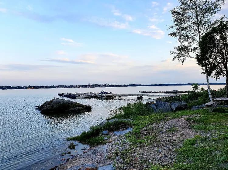 En klarblå himmel speglas i det stilla vattnet vid Gräsöbadens Familjecamping, omgiven av naturliga klippformationer och grönskande landskap vid horisonten.