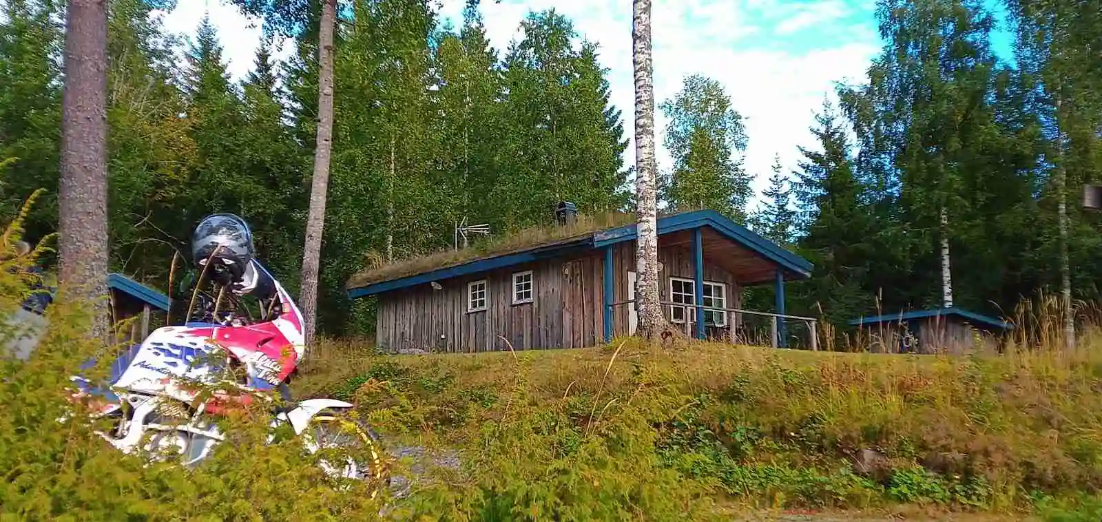 En motorcykel står parkerad framför en rustik timmerstuga omgiven av grönskande skog.