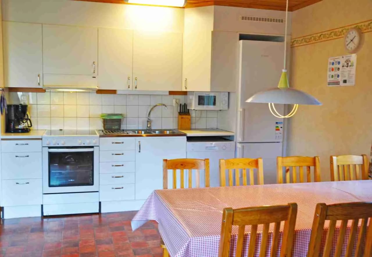 Bild av ett modernt kök med trädetaljer. Köket har ljusa träskåp och en mörk bänkskiva. En stor vitvaraa står vid sidan och golvet är av trä. Takhöjden och övriga inredningen bidrar till en ljus och rymlig atmosfär.