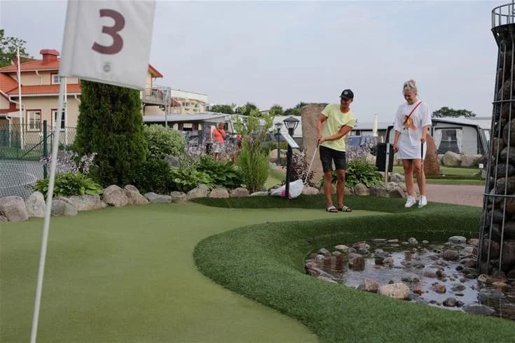 Personer spelar minigolf på en gräsmatta omgiven av lummig grönska vid Grebbestadfjorden. Golfklubbor och -utrustning syns tydligt, vilket bidrar till en trivsam och avkopplande fritidsmiljö.