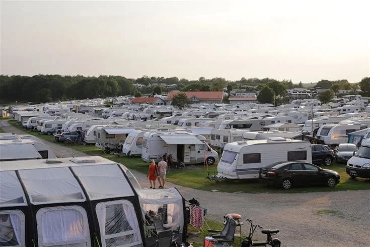 En campingplats vid Grebbestadfjorden med flera husbilar och husvagnar parkerade på en stor parkering, omgiven av grönska och träd i bakgrunden.