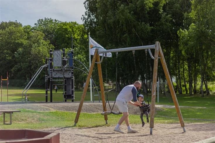 En lekplats i Grebbestadfjorden omgiven av träd och grönska, med människor som njuter av utomhusaktiviteter i en parkmiljö.