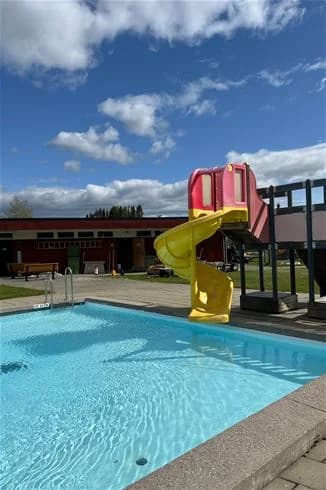 En campinganläggning med en utomhuspool där människor simmar och leker under en solig dag. I bakgrunden syns grönska och campingens byggnader.