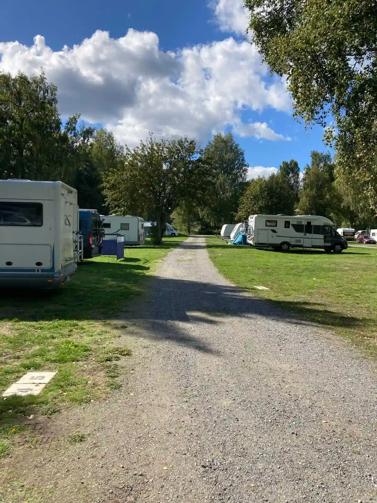 En husvagn och en lastbil står parkerade i en skogsområdets parkeringsplats nära Gålö havsbad. Träd omger fordonen, vilket ger en naturnära inramning.