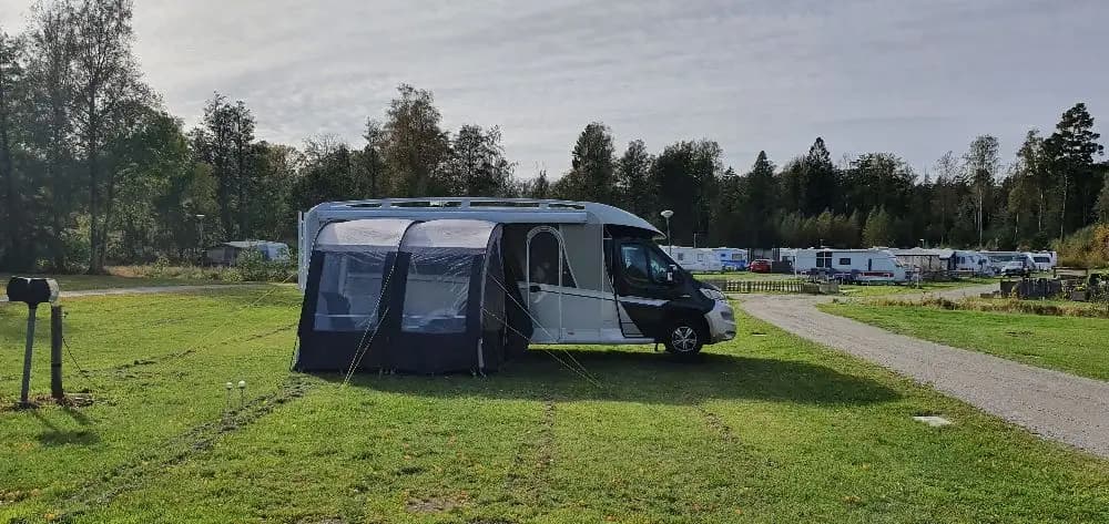 En campingplats på Gålö havsbad med uppställda husbilar och tält, omgivna av grönska och parkerade fordon. En solig dag med klar himmel skapar en avslappnad atmosfär för camping och fritidsaktiviteter.
