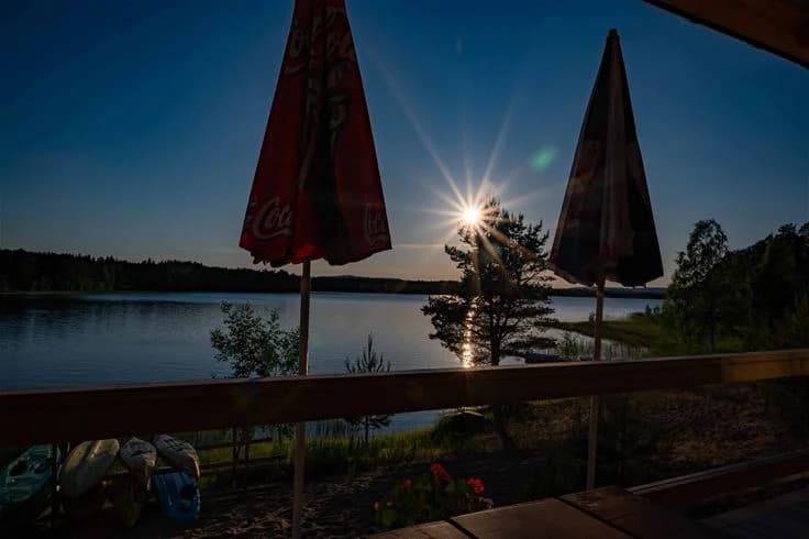 En naturskön kvällsbild över Norra Dellen Familjecamping med en spegelblank sjö i förgrunden som reflekterar solnedgångens varma färger. Utemöbler syns vid strandkanten, vilket skapar en inbjudande atmosfär för besökare att njuta av den stilla omgivningen.