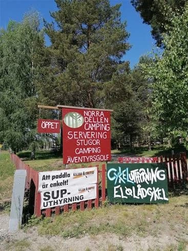 En skylt vid Norra Dellen Familjecamping omgiven av grönskande bladverk i morgonljus.