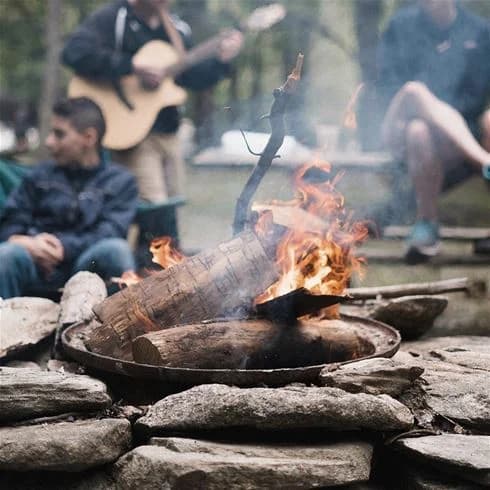 En grupp människor sitter runt en lägereld vid Storforsens camping. Elden flammar livligt och sprider värme och glöd medan skymningen sänker sig över naturen.