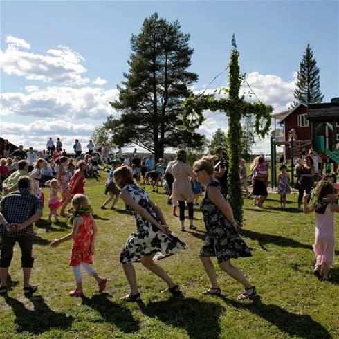 En livlig campingplats med många besökare som njuter av en solig dag vid Storforsen. Människor samlas för aktiviteter och rekreation, medan ett evenemang med stänger och dekorativa inslag pågår i bakgrunden. Stämningen är avslappnad och underhållande, med en stark känsla av gemenskap.