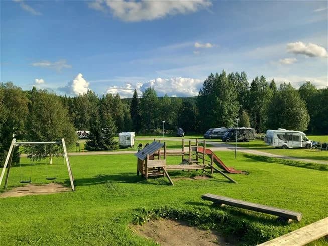 En grönskande campingplats vid Ristafallet omgiven av träd och öppna ängar under en molnig himmel.