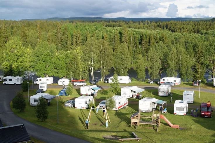 En fågelperspektivbild över Ristafallets camping visar ett vackert naturlandskap med frodiga träd, gröna ängar och uppställda husbilar. Området omges av täta barrträd, vilket ger en känsla av avskildhet och närhet till naturen.