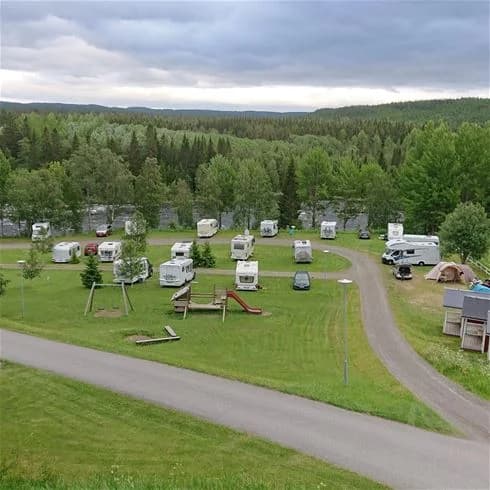 En campingplats vid Ristafallets camping med gräsbevuxna ytor, omgivande växter och en husbil parkerad i en naturskön miljö.