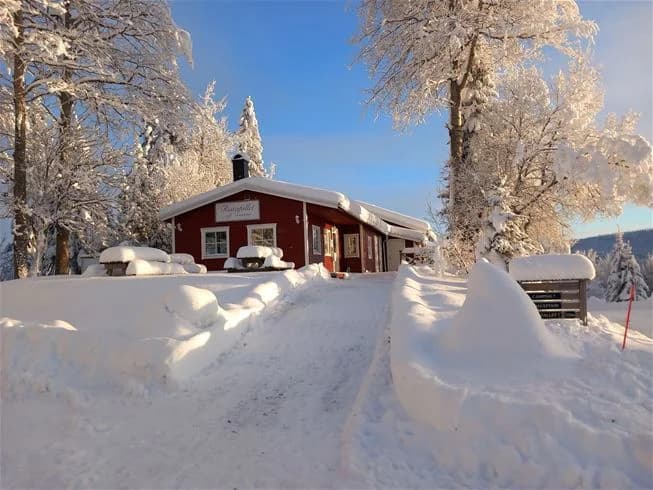 En snötäckt campingplats vid Ristafallet, med träd omgivna av vinterlandskap. I bakgrunden syns ett litet hus eller stuga täckt av snö.