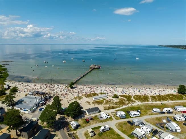 Flygfoto av Köpingbaden Camping med en naturskön kustlinje. Sandstrand som vetter mot ett stort hav, omgiven av grönskande landskap. Perfekt för strandaktiviteter och avkoppling vid vattnet.