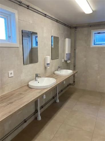 En ren och modern badrumsanläggning med vita väggar och golv. Ett handfat med kran samt ett spegelstänkskydd syns i bild. Tecken på en funktionell och genomtänkt inredning för besökare.