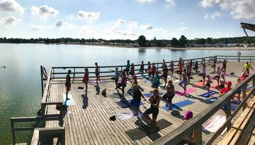 En bild av Köpingbaden Camping med semesterfirare som promenerar längs en träbrygga med vy över vattnet. Området är aktivt med fritidsaktiviteter och är en populär turistattraktion.