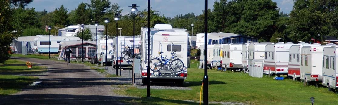 En husbil parkerad på en campingplats omgiven av grönska, med andra campare och fordon synliga i bakgrunden.