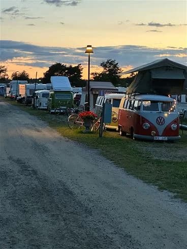 En campingplats med husbilar och tält uppställda på en grusparkering under en delvis molnig himmel. Fritidsfordon och en transportbil syns i bakgrunden.