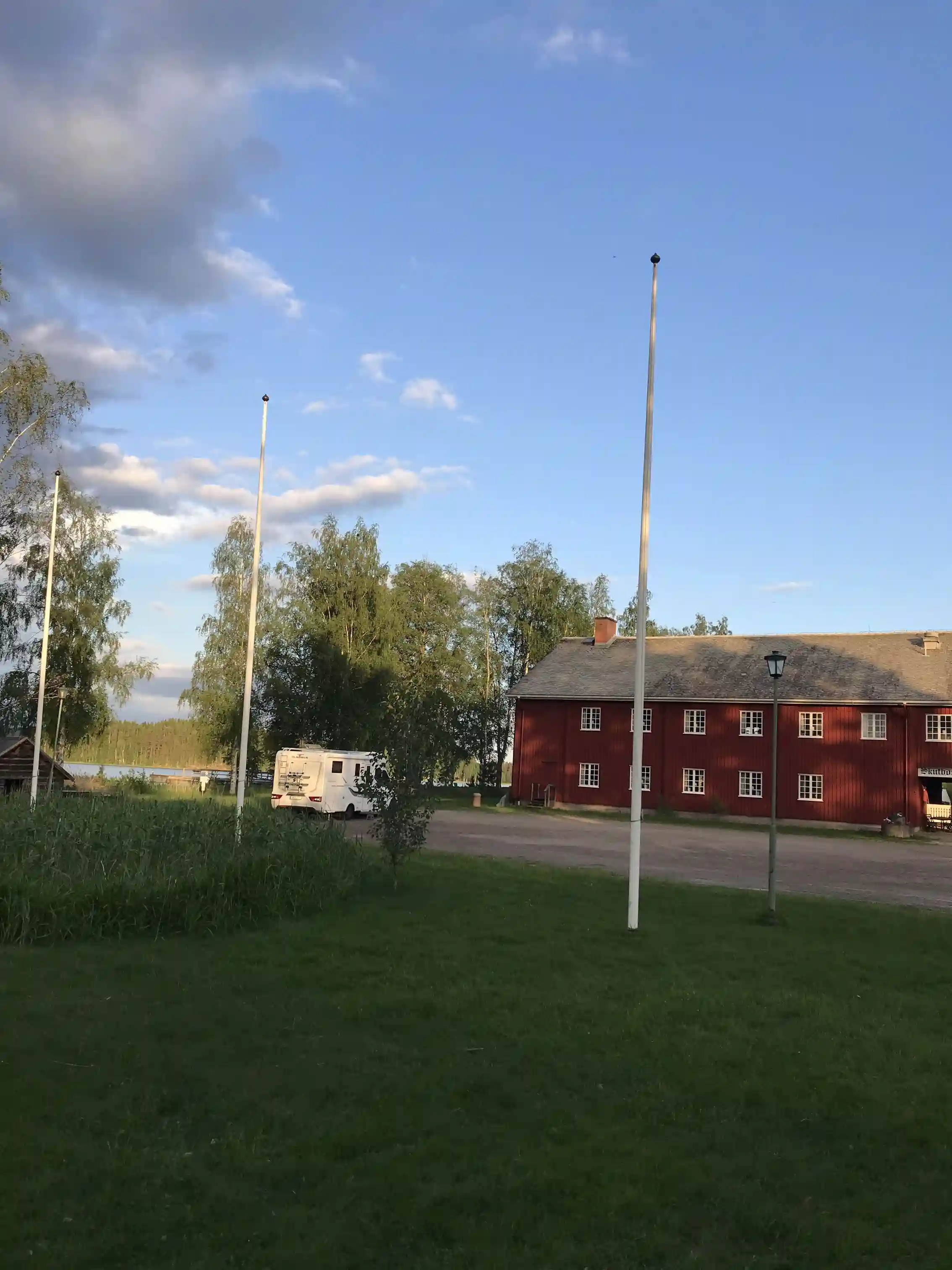 Bild som visar en öppen gräsyta med en historisk byggnad vid Brunskog hembygdsgård. Det är kväll och en gatulampa lyser upp området vars omgivning inkluderar en asfalterad uppfart och en landsväg.