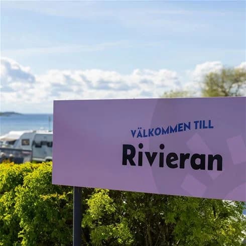 En campingplats vid vattnet med båtar förtöjda vid en brygga, omgiven av grönska och klara blå himmel.