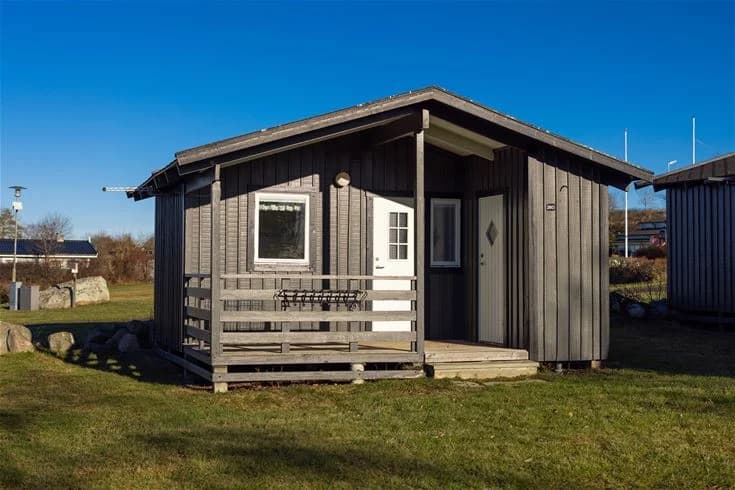 En liten trästuga med rött tak och vita detaljer står på en grön gräsmatta på Klinta Camping. En enkel trädörr och fönster syns på stugans fasad.