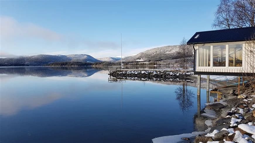 En vinterbild av Jormvattnets Fiskecamp med en snötäckt sjö och omgivande landskap, vars yta speglar den klara vinterhimlen.