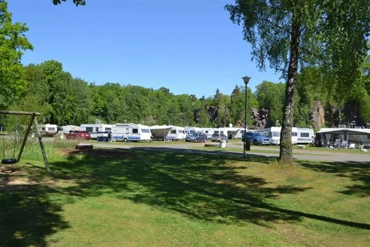 En campingplats med träd och grönska, där husvagnar och bilar är parkerade. Området har en öppen gräsyta och skuggiga platser under träden. Platsen förefaller vara en trevlig miljö för fritidsaktiviteter och rekreation.