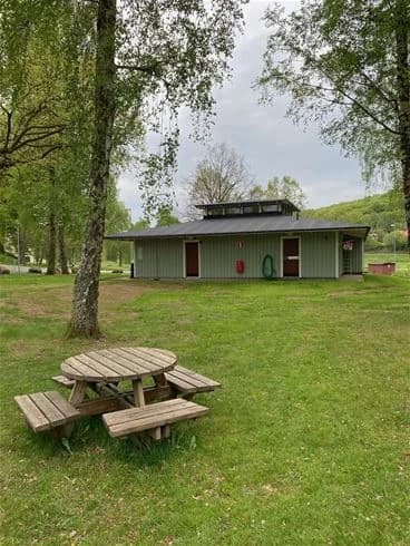 En öppen gräsyta med ett lantligt hus i bakgrunden, omgiven av träd och lågväxt vegetation.