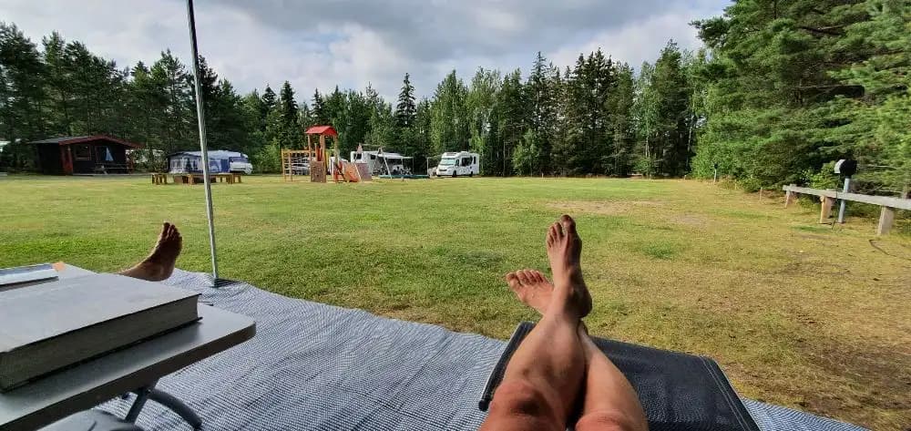 En grön gräsmatta omgiven av träd där flera campingtält står uppställda. Utemöbler syns i förgrunden och människor njuter av fritiden i parkliknande miljö på Habo Camping och Stugby.