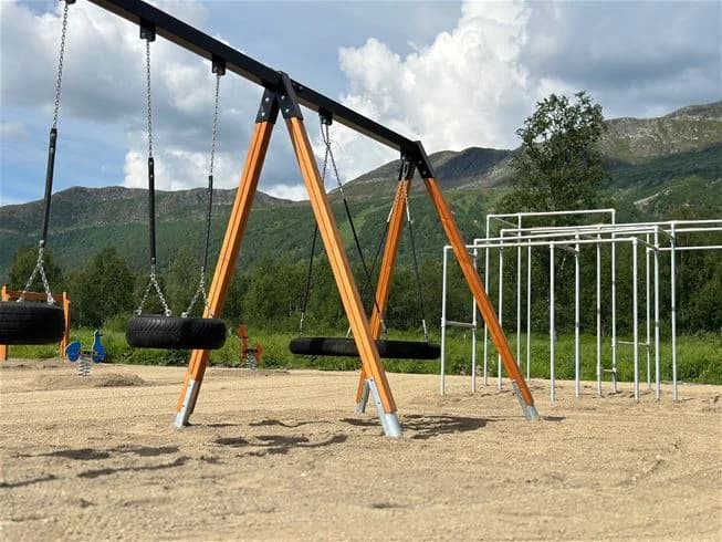 En gunga och balansutrustning i en utomhuslekplats omgiven av sand och rep med bakgrund av bebyggelse och parkmiljö.