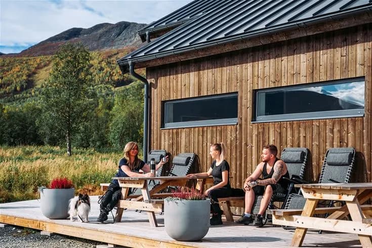 En timrad stuga med träfasad och sittplatser utomhus vid ett picknickbord, omgiven av grönskande natur i Camp Kittelfjäll.