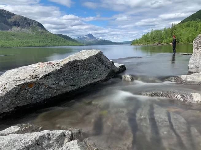 En storslagen vy över bergigt landskap vid Camp Kittelfjäll, med en klar vattenyta som reflekterar de omgivande bergen. I bakgrunden syns moln svepa över bergstopparna.