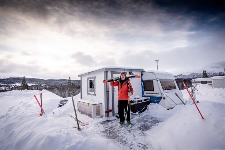En snötäckt vinterlandskap med en parkerad skoter bredvid ett litet tält eller stuga i Camp Kittelfjäll. Bakom syns en vidsträckt is- och snöscen med små snödrivor och frostiga träd.