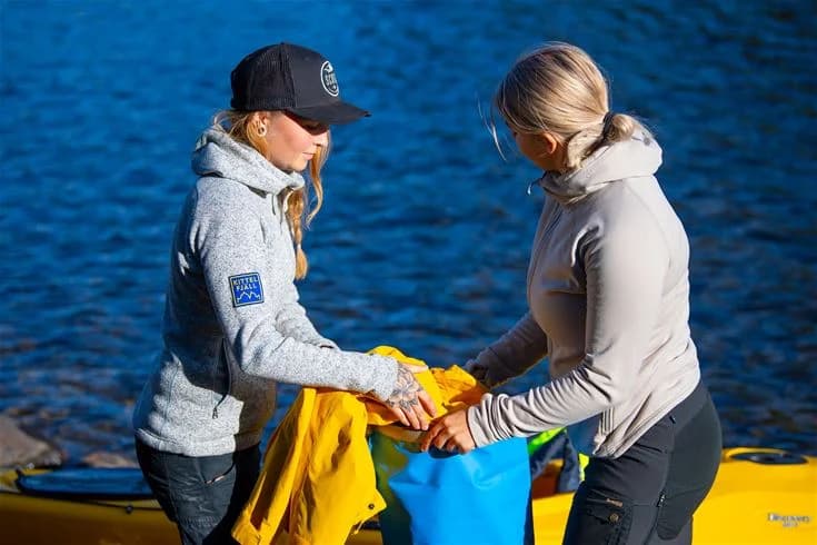 En person klädd i en jacka och skyddsglasögon njuter av en solig dag vid havet, omgiven av vågor. Det finns en känsla av avkoppling och semester.