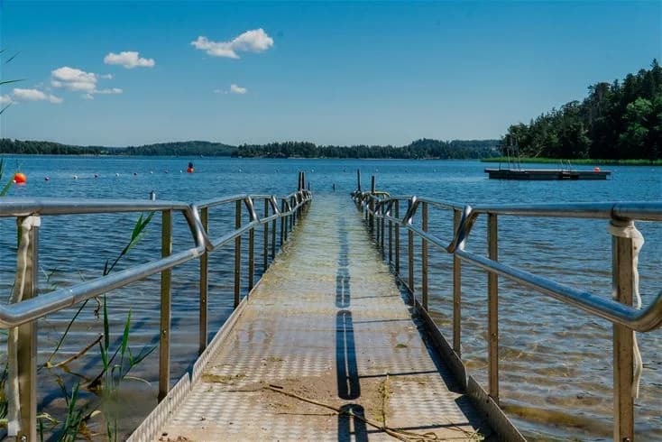 En solig dag vid Farstanäs camping & havsbad med en brygga som sträcker sig ut i det glittrande vattnet, omgiven av sjö- och kustlandskap.