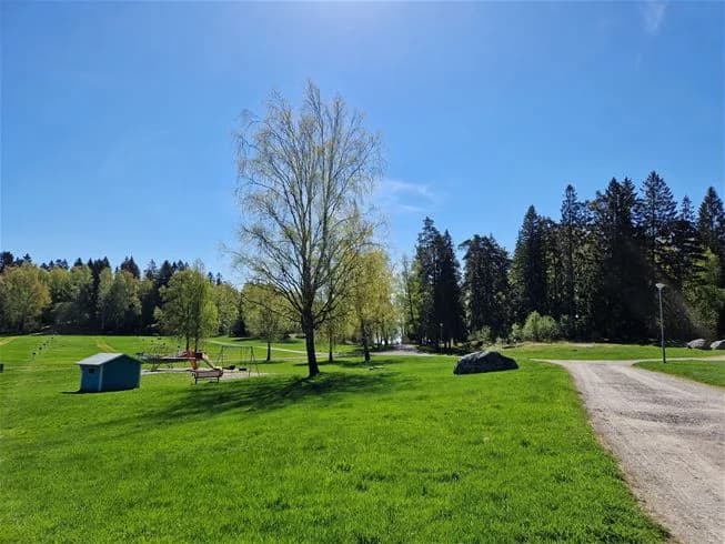 En vidsträckt gräsplan på Farstanäs Camping & Havsbad med flera träd i bakgrunden under en molnig himmel.