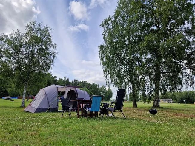 En tältplats på Farstanäs Camping & Havsbad, omgiven av grönskande växter och träd. Ett tält står uppställt på en välskött gräsmatta, delvis i skugga, vilket skapar en avslappnad campingmiljö i parkliknande natur.