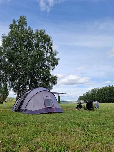 En vidsträckt campingplats vid Farstanäs camping & havsbad. I förgrunden syns ett tält uppställt på en grön äng, omgiven av öppen mark och några träd i bakgrunden. Utrustning för friluftsliv ligger vid tältet, vilket ger en känsla av avkoppling nära naturen.