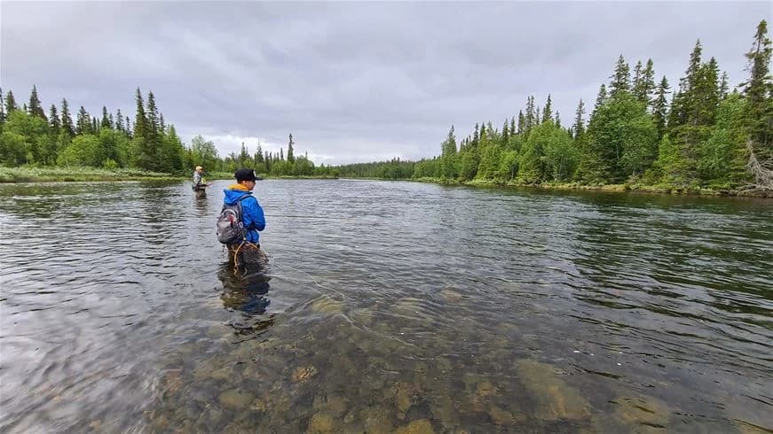 En avkopplande scen vid Jormvattnets Fiskecamp med en fiskare vid kanten av en flod eller sjö omgiven av vildmark och molntäckta himlar.