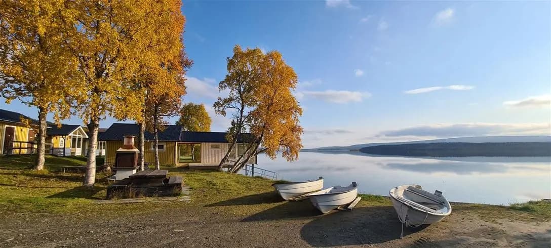 En lugn sjö med en röd stuga vid stranden, omgiven av skogslandskap. Flera små båtar ligger förtöjda vid vattnet under en klar himmel vid Jormvattnets fiskecamp.