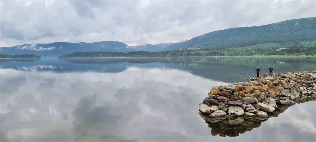 Vy över Jormvattnets fiskecamp med ett glittrande vattenlandskap. I förgrunden syns en lugn sjö, omgiven av berg och klippor. Moln svävar över de bergiga landformerna i bakgrunden, vilket skapar en naturskön och avkopplande atmosfär.
